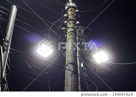 Concrete utility pole illuminated by bright LED street lamps at night, tangled power lines crisscrossing the dark sky, high-contrast urban scenery capturing industrial infrastructure during late hours Concrete utility pole illuminated by bright LED street lamps at night, tangled power lines crisscrossing the dark sky, high-contrast urban scenery capturing industrial infrastructure during late hours 140041929