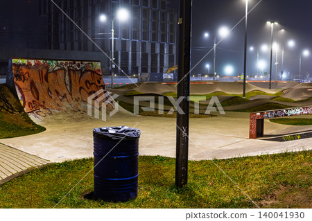 A lonely blue metal trash bin stands in a deserted urban skatepark at night, illuminated by bright glowing street lamps under a misty winter sky A lonely blue metal trash bin stands in a deserted urban skatepark at night, illuminated by bright glowing street lamps under a misty winter sky 140041930