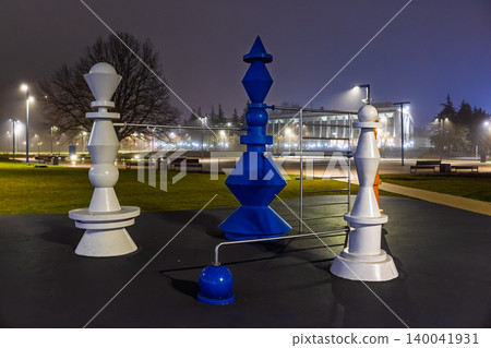 Large decorative chess pieces installation in a public park at night, featuring stylized blue and white figures linked by metal bars under glowing streetlights against a dim urban background Large decorative chess pieces installation in a public park at night, featuring stylized blue and white figures linked by metal bars under glowing streetlights against a dim urban background 140041931