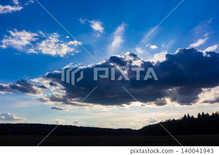 Dramatic sunbeams piercing through heavy dark clouds in a vibrant blue sky over a serene countryside horizon, creating a striking atmosphere of impending weather and natural beauty Dramatic sunbeams piercing through heavy dark clouds in a vibrant blue sky over a serene countryside horizon, creating a striking atmosphere of impending weather and natural beauty 140041943