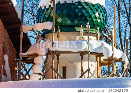 Intricate green onion dome of a Russian Orthodox church, actively under renovation. Winter snow blankets scaffolding against a crisp blue sky, Russia 140042179