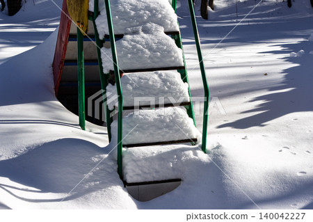 Fresh powder blankets a vibrant green slide ladder, creating a winter wonderland vibe. Sunlight highlights crisp shadows on pure white snow 140042227