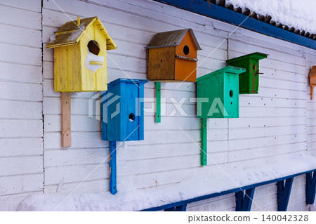 A row of brightly painted birdhouses hangs against a white wooden wall under a snowy eave, offering a colorful contrast to the winter scene 140042328