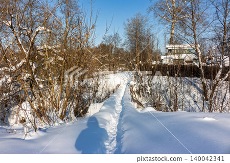 Crisp winter day. A serene, snow-covered trail winds over a gentle ravine, beckoning explorers through a quiet, frosty landscape under a bright blue sky 140042341