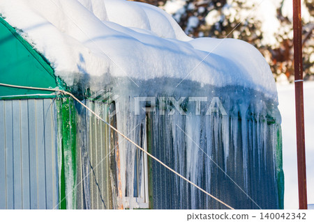 Frosty construction dwelling under winter's grip. Roof piled high with snow, gleaming sharp icicles adorn the eaves in bright sunlight Frosty construction dwelling under winter's grip. Roof piled high with snow, gleaming sharp icicles adorn the eaves in bright sunlight 140042342