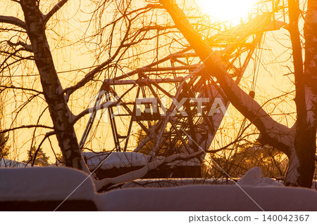 Massive industrial crane's steel frame silhouetted by a dramatic winter sunset. Golden light glows through bare tree branches, creating a powerful, frozen scene Massive industrial crane's steel frame silhouetted by a dramatic winter sunset. Golden light glows through bare tree branches, creating a powerful, frozen scene 140042367