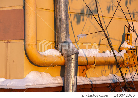 Glistening icicles dangle on a bare branch, catching the golden evening light against snow-dusted pipes and a warm yellow wall, signaling the grip of winter 140042369