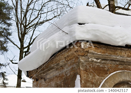 Thick snow overhangs an old, weathered brick cornice under a pale winter sky, bare tree branches visible in background Thick snow overhangs an old, weathered brick cornice under a pale winter sky, bare tree branches visible in background 140042517