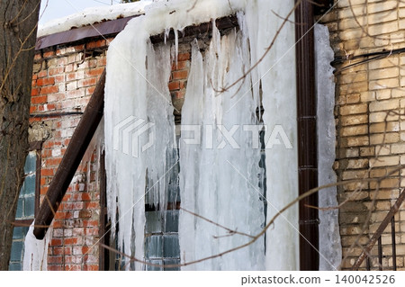 Massive icicles hang like frozen curtains from the building eaves, contrasting with the weathered brick facade and brown drainpipe Massive icicles hang like frozen curtains from the building eaves, contrasting with the weathered brick facade and brown drainpipe 140042526