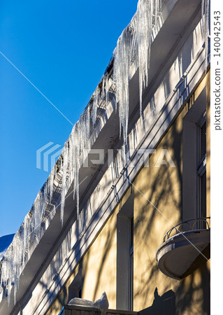 Long sharp icicles dangle from a building cornice against a brilliant blue sky, dripping in a winter thaw bright sunlight Long sharp icicles dangle from a building cornice against a brilliant blue sky, dripping in a winter thaw bright sunlight 140042543
