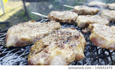 Pork steaks sizzle on home grill as the surface slowly browns. Smoke rises gently into the air while the meat cooks evenly over steady heat, capturing the rich atmosphere of relaxed backyard barbecue 140042879