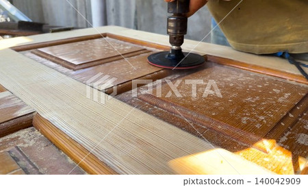 Male hands use electric polishing machine to smooth and refine wooden door surface. The rotating pad carefully treats wood, enhancing texture and finish during detailed restoration or renovation work 140042909