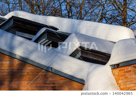 Thick blankets of winter snow drape over a pitched roof with dormer windows, contrasting against a bright blue sky and bare tree branches Thick blankets of winter snow drape over a pitched roof with dormer windows, contrasting against a bright blue sky and bare tree branches 140042965