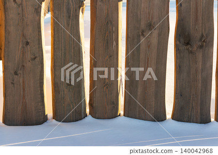 Weathered wooden fence panels stand tall against bright snow, with warm sunlight backlighting the rough-hewn edges for a striking contrast in the winter landscape Weathered wooden fence panels stand tall against bright snow, with warm sunlight backlighting the rough-hewn edges for a striking contrast in the winter landscape 140042968