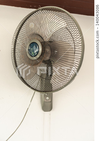 Wall mounted electric fan with metal grille on white interior wall, household cooling device for air circulation during hot weather Wall mounted electric fan with metal grille on white interior wall, household cooling device for air circulation during hot weather 140043086