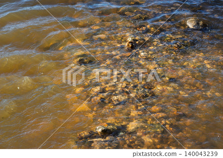 Clear river water flows over a bed of smooth colorful pebbles at the shore, creating gentle ripples and light reflections on the submerged stones under the summer sun Clear river water flows over a bed of smooth colorful pebbles at the shore, creating gentle ripples and light reflections on the submerged stones under the summer sun 140043239