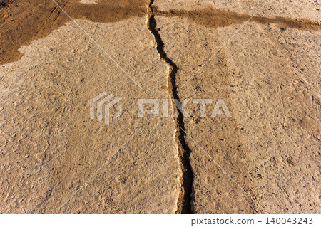 Deep jagged crack splitting an old weathered concrete road surface, showing textured gray cement and dry dusty earth in a remote countryside location under soft natural daylight Deep jagged crack splitting an old weathered concrete road surface, showing textured gray cement and dry dusty earth in a remote countryside location under soft natural daylight 140043243