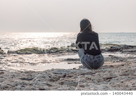 The back view of a woman quietly gazing at the sea from a rocky beach. 140043440