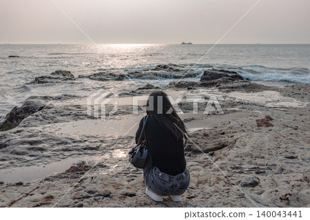 The back view of a woman quietly gazing at the sea from a rocky beach. 140043441