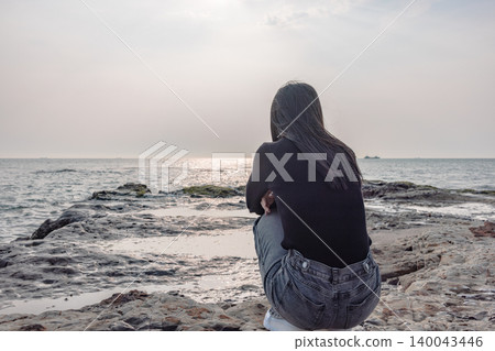 The back view of a woman quietly gazing at the sea from a rocky beach. The back view of a woman quietly gazing at the sea from a rocky beach. 140043446