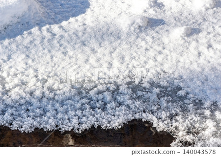 Brilliant white frost crystals meet dark water at the edge of a frozen stream, a crisp winter morning close-up, sunlit detail texture 140043578