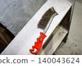 A well-worn metal wire brush with a bright orange handle rests on a distressed white wooden shelf, suggesting renovation or cleaning tasks 140043624