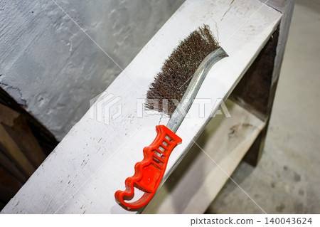 A well-worn metal wire brush with a bright orange handle rests on a distressed white wooden shelf, suggesting renovation or cleaning tasks A well-worn metal wire brush with a bright orange handle rests on a distressed white wooden shelf, suggesting renovation or cleaning tasks 140043624