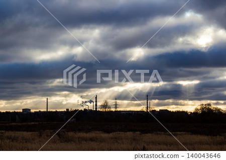 Dramatic sky with sun rays piercing through heavy clouds over an industrial landscape silhouette. Smokestacks puffing against the sunset Dramatic sky with sun rays piercing through heavy clouds over an industrial landscape silhouette. Smokestacks puffing against the sunset 140043646