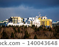 Vibrant modern residential complex peeking over a wintry forest, showcasing urban development against a dramatic blue sky. Contemporary housing meets nature's edge 140043648