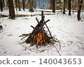 A cozy bonfire blazes warmly in a serene, snow-covered forest, its orange glow fighting the winter chill. Perfect for cold season adventures 140043653