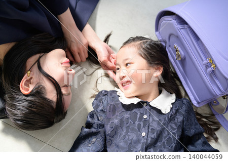 A Japanese parent and child are lying on the floor, smiling and gazing at each other with their school bags in hand. A Japanese parent and child are lying on the floor, smiling and gazing at each other with their school bags in hand. 140044059