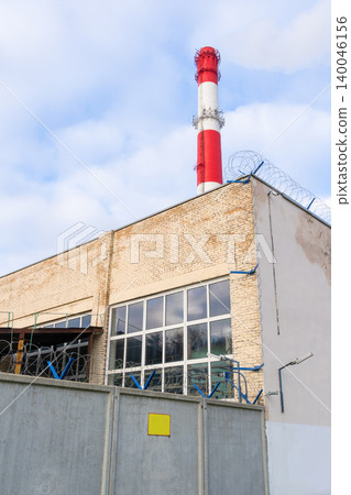 Factory chimney under bright daytime. Clear sky highlights towering chimney beside factory windows Factory chimney under bright daytime. Clear sky highlights towering chimney beside factory windows 140046156