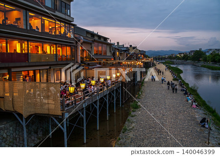 A photograph of Kyoto's scenery at dusk, with lights lit up on the Kamo River and riverside terraces. 140046399
