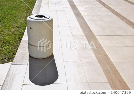 A cylindrical trash bin made of concrete stands on a tiled pavement, surrounded by green grass. The bin features a circular opening and a clean, minimalist design in a sunny outdoor environment A cylindrical trash bin made of concrete stands on a tiled pavement, surrounded by green grass. The bin features a circular opening and a clean, minimalist design in a sunny outdoor environment 140047399