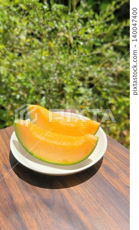 A fresh, juicy red-fleshed cut melon on a plate, photographed from a diagonal angle above against a backdrop of lush greenery. A fresh, juicy red-fleshed cut melon on a plate, photographed from a diagonal angle above against a backdrop of lush greenery. 140047400