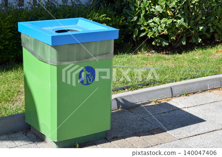A green recycling bin with a blue lid is positioned on a paved sidewalk next to a grassy area, featuring a symbol indicating its purpose for waste disposal and recycling A green recycling bin with a blue lid is positioned on a paved sidewalk next to a grassy area, featuring a symbol indicating its purpose for waste disposal and recycling 140047406