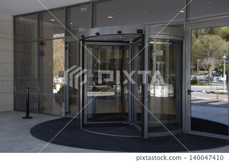 A sleek, modern glass revolving door is prominently featured at the entrance of a commercial building, surrounded by large glass panels and a clean, minimalistic interior design A sleek, modern glass revolving door is prominently featured at the entrance of a commercial building, surrounded by large glass panels and a clean, minimalistic interior design 140047410