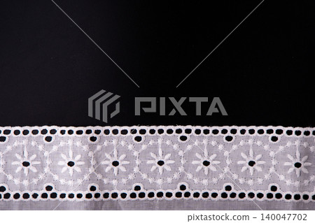 White floral lace ribbon trim on black background, delicate cotton embroidery border fabric mockup. Top view embroidered eyelet flower patterns on a black surface. Text copy space template White floral lace ribbon trim on black background, delicate cotton embroidery border fabric mockup. Top view embroidered eyelet flower patterns on a black surface. Text copy space template 140047702