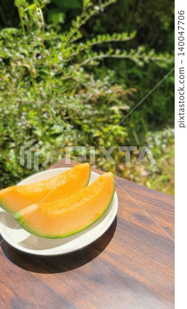 Fresh, juicy red-fleshed cut melons arranged on a white plate, photographed outdoors amidst the beautiful fresh greenery. Fresh, juicy red-fleshed cut melons arranged on a white plate, photographed outdoors amidst the beautiful fresh greenery. 140047706
