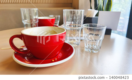 Empty red coffee cup and clear drinking glasses on a cozy cafe table in soft daylight, creating a warm and relaxing atmosphere for breakfast or informal meetings. Empty red coffee cup and clear drinking glasses on a cozy cafe table in soft daylight, creating a warm and relaxing atmosphere for breakfast or informal meetings. 140048423