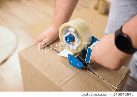 Close-up of man hands using tape gun dispenser to seal cardboard box for moving. 140048945