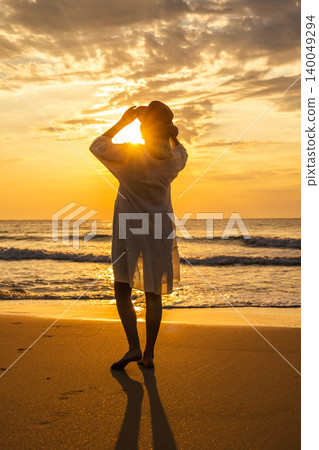 Middle aged woman on beach at sunrise adjusting hat, peaceful freedom travel lifestyle by sea 140049294