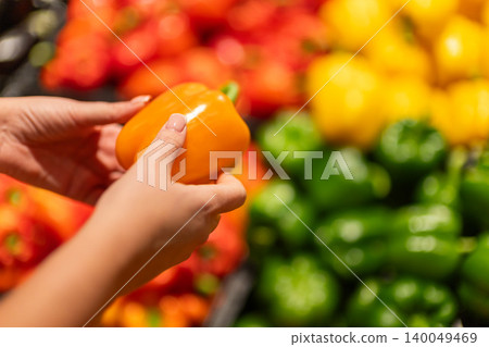 Woman hands holding fresh orange bell pepper in supermarket produce section, colorful vegetables background for healthy grocery shopping concept Woman hands holding fresh orange bell pepper in supermarket produce section, colorful vegetables background for healthy grocery shopping concept 140049469