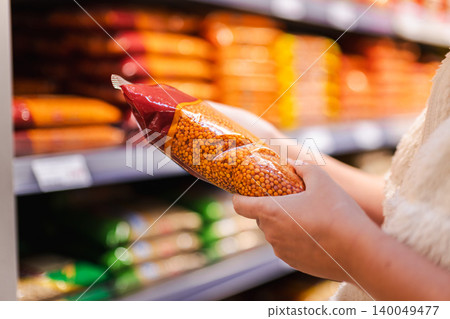 Woman choosing packaged red lentils in supermarket aisle, customer holding dry legumes product while shopping for healthy grocery ingredients 140049477