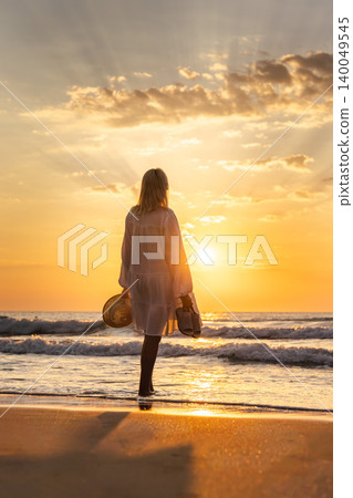 Middle aged woman standing on beach at sunrise holding hat and shoes, freedom travel lifestyle by sea Middle aged woman standing on beach at sunrise holding hat and shoes, freedom travel lifestyle by sea 140049545