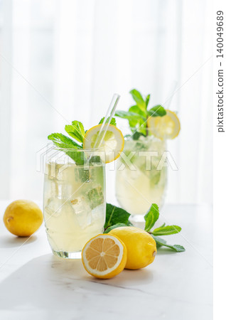 A summer drink with lemon, mint, and ice in a two glasses against a sunny window background. A summer drink with lemon, mint, and ice in a two glasses against a sunny window background. 140049689
