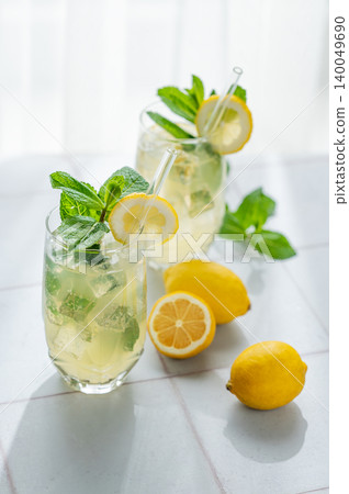 A summer drink with lemon, mint, and ice in a two glasses against a sunny window background. A summer drink with lemon, mint, and ice in a two glasses against a sunny window background. 140049690