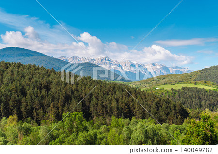 Panoramic view of Pyrenees mountains in Catalonia with dense forest green valley and snow capped peaks under blue sky Panoramic view of Pyrenees mountains in Catalonia with dense forest green valley and snow capped peaks under blue sky 140049782