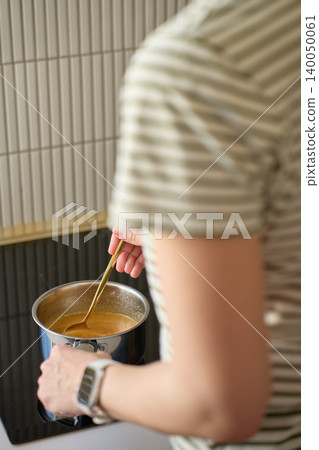 Woman stirs mixture in metal saucepan on kitchen stovetop 140050061