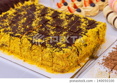 Dessert with yellow crumbs, brown icing is cut into squares on table for celebration with other treats nearby. Dessert with yellow crumbs, brown icing is cut into squares on table for celebration with other treats nearby. 140050100